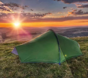 Green backpacking tent pitched on a grassy hilltop overlooking rolling hills and valleys at sunrise, the sun's golden rays and colourful clouds lighting the sky.