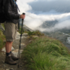 Partial view of a hiker's legs and hands holding a trekking pole on a rocky mountain path, wearing hiking boots, shorts and a jacket, overlooking a cloud-filled valley with a winding river and wind-swept grasses in the foreground