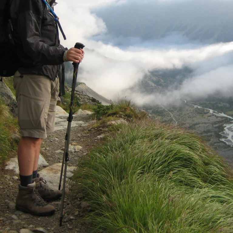 Partial view of a hiker's legs and hands holding a trekking pole on a rocky mountain path, wearing hiking boots, shorts and a jacket, overlooking a cloud-filled valley with a winding river and wind-swept grasses in the foreground