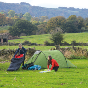 Two people setting up camp beside a green tent in a grassy field—one kneeling in a red jumper arranging a sleeping mat and the other in a navy hoodie holding a black-and-red sleeping bag—with a dry-stone wall and tree-covered rolling hills in autumn colours behind them.
