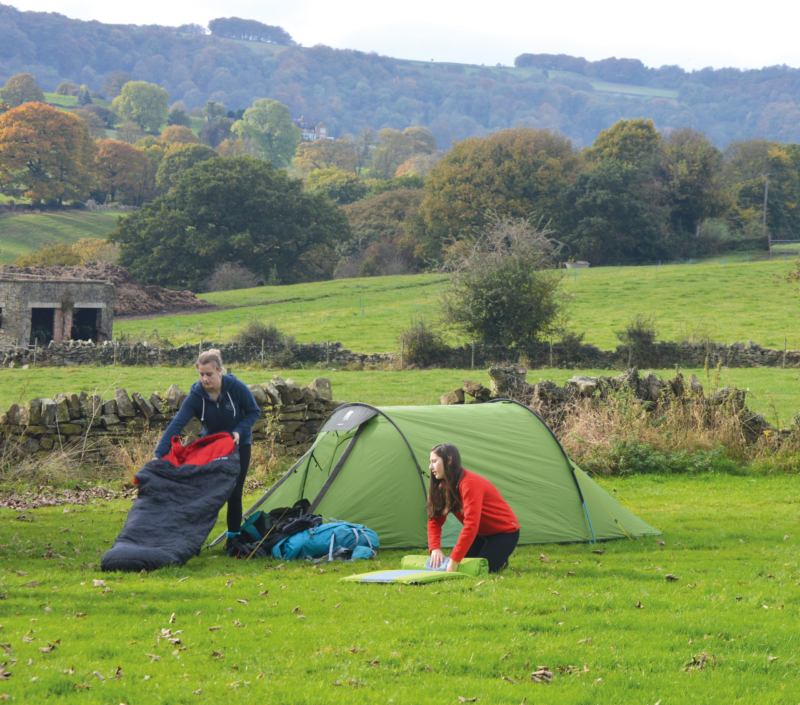 Two people setting up camp beside a green tent in a grassy field—one kneeling in a red jumper arranging a sleeping mat and the other in a navy hoodie holding a black-and-red sleeping bag—with a dry-stone wall and tree-covered rolling hills in autumn colours behind them.