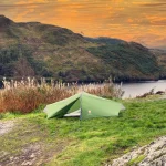 Green two-person tent pitched on a grassy rocky ridge beside tall dried wildflowers, overlooking a calm lake and tree-covered hills beneath an orange sunset sky.