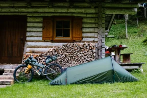 Small green backpacking tent pitched on grass beside a rustic log cabin with a stacked pile of firewood, a fully loaded touring bicycle leaning against the woodpile and a wooden picnic table with cycling helmets in a forest clearing.