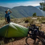 Cyclist in a blue jersey drinks from a water bottle beside a small green tent and a loaded touring bike on a sunlit mountain ridge with forested hills and pine trees beyond.