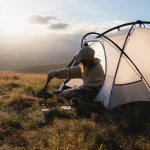 Person in a cap and fleece sitting at the entrance of a light-coloured tent on a grassy hillside, stirring a small frying pan on a portable camping stove as golden sunlight falls over rolling hills.