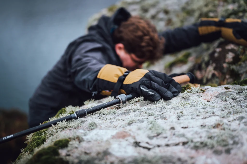 ls_ex_Capitol_Peak-2022 A climber's gloved hand (black and yellow, 'extremities' logo visible) grips a frost-dusted rocky ledge beside a trekking pole, the climber blurred in the background.