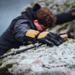 A climber's gloved hand (black and yellow, 'extremities' logo visible) grips a frost-dusted rocky ledge beside a trekking pole, the climber blurred in the background.