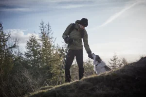 Hiker in a green puffer jacket and black beanie standing on a grassy hill with trekking poles, bending to pet a fluffy white-and-grey dog against a backdrop of conifer trees and a pale sky.