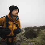 Hiker in a mustard-yellow jacket, black beanie and gloves carrying a backpack walks a rocky path across foggy moorland while looking up into the misty distance.