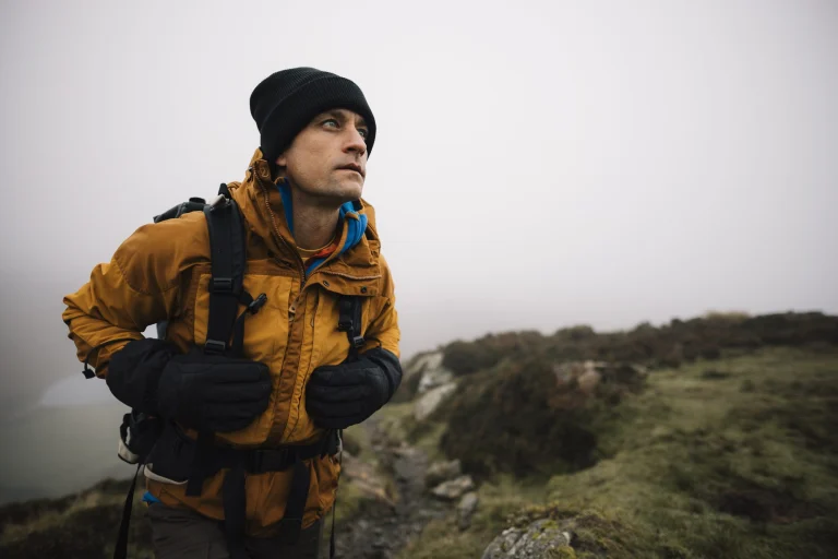 Hiker in a mustard-yellow jacket, black beanie and gloves carrying a backpack walks a rocky path across foggy moorland while looking up into the misty distance.