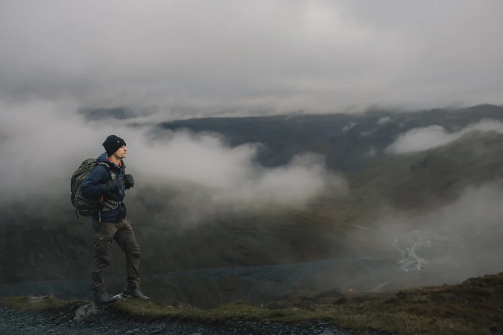 A lone hiker in a dark beanie, gloves and backpack stands on a grassy ridge looking out over a mist-filled valley and rolling hills beneath a low grey sky.