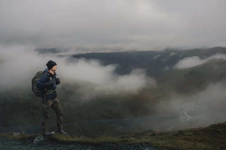 A lone hiker in a dark beanie, gloves and backpack stands on a grassy ridge looking out over a mist-filled valley and rolling hills beneath a low grey sky.