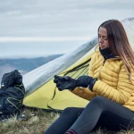 A woman in a yellow insulated jacket sits outside a lightweight tent on a grassy hillside, pulling on black gloves beside a backpack with distant rolling hills under a cloudy sky.