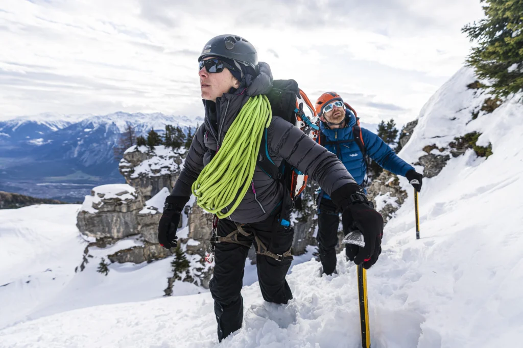 Two alpine climbers ascending a steep snowy slope — the lead climber in a black helmet and jacket carries a bright green climbing rope over their shoulder while a second climber in a blue jacket and orange helmet follows with ice axes, rocky snow-covered peaks visible behind them.