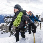Two alpine climbers ascending a steep snowy slope — the lead climber in a black helmet and jacket carries a bright green climbing rope over their shoulder while a second climber in a blue jacket and orange helmet follows with ice axes, rocky snow-covered peaks visible behind them.