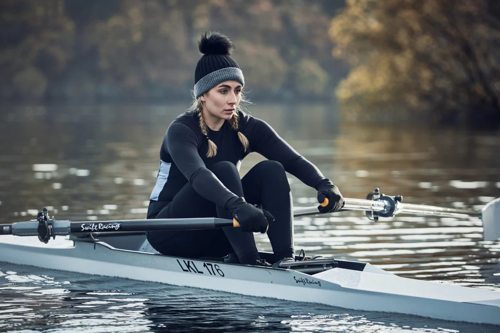 ls_ex_kyak_LIN-TERRANOVA-2022 Woman in a black thermal outfit and grey-and-black pom-pom beanie with braided hair, pulling oars on a single scull across a calm lake with blurred autumn trees behind her