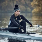 Woman in a black thermal outfit and grey-and-black pom-pom beanie with braided hair, pulling oars on a single scull across a calm lake with blurred autumn trees behind her