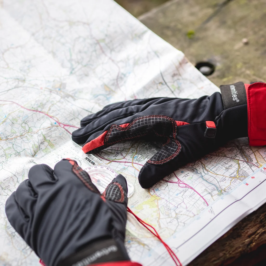 Pair of black and red outdoor gloves resting on an unfolded topographic map, one glove showing textured palm stitching and the other near a clear baseplate compass with a red lanyard on a wooden bench.