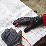 Pair of black and red outdoor gloves resting on an unfolded topographic map, one glove showing textured palm stitching and the other near a clear baseplate compass with a red lanyard on a wooden bench.