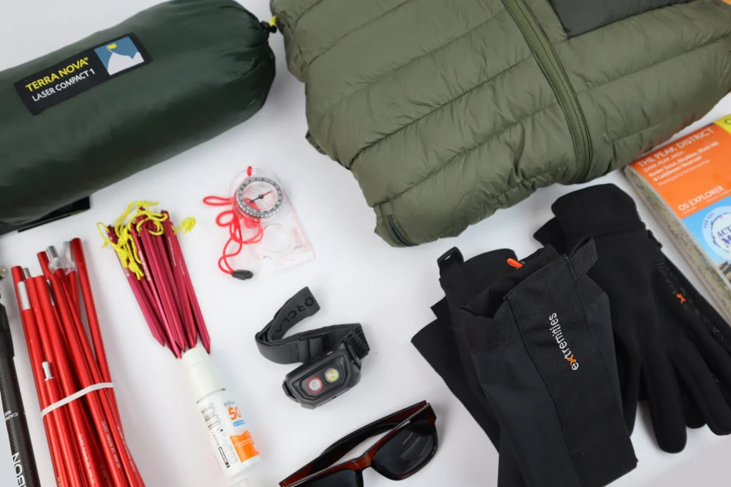 ls_flatlay_1_2024 Flat-lay of camping gear on a white background showing a green Terra Nova Laser Compact 1 sleeping bag in its stuff sack, an olive quilted jacket, black gloves, sunglasses, a headlamp, a clear-base compass with red cord, bundles of red tent poles and pegs, a bottle of SPF50 sunscreen and folded maps.