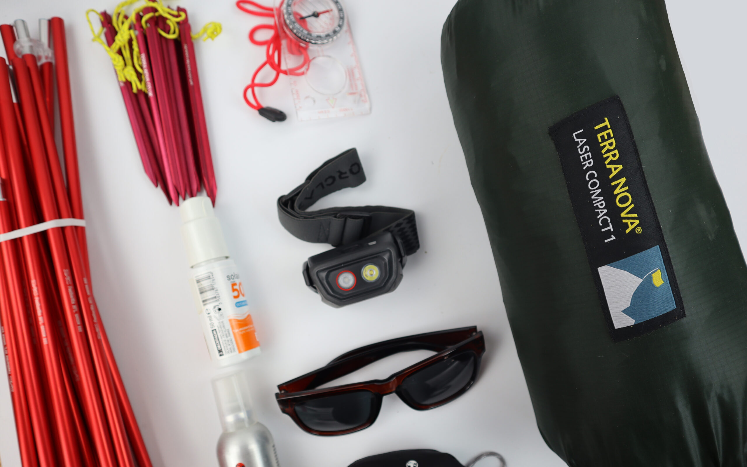 Flat-lay of camping gear on a white background: red aluminium tent poles and red pegs with yellow guy-lines, a compact green Terra Nova Laser Compact 1 tent bag, a black headlamp, sunglasses, a compass, a bottle of sunscreen and a small gas canister.
