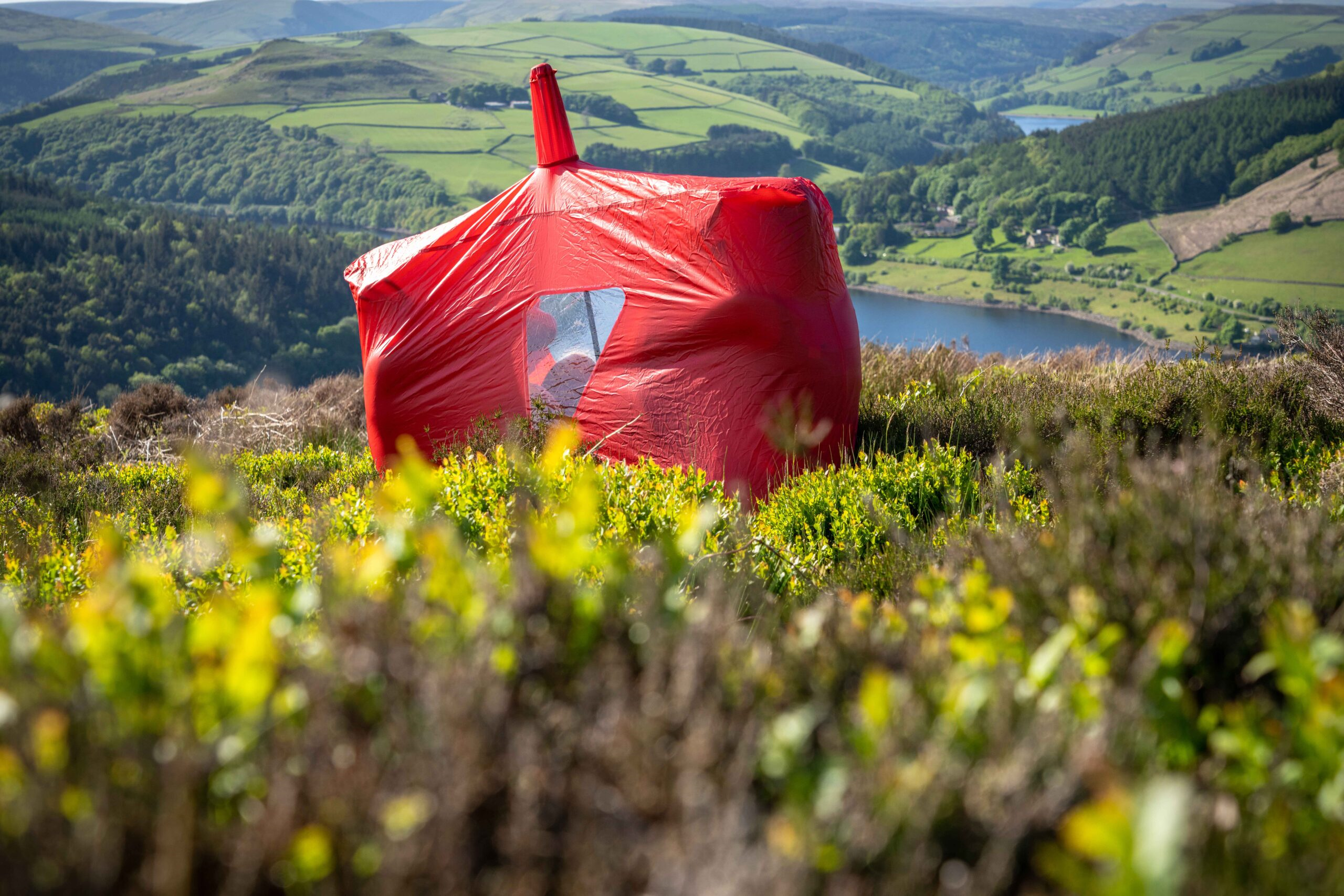 Bright red bivvy-style shelter with a small clear window pitched in green heather on a hillside, overlooking a reservoir, patchwork fields and wooded valleys.