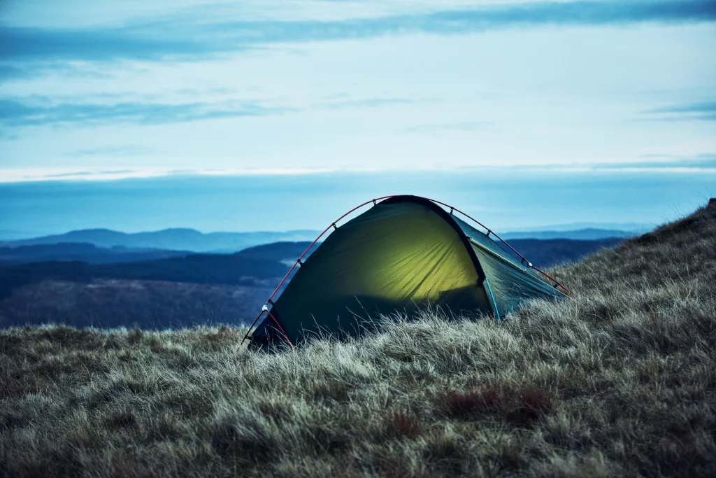 ls_moor_twilight_Southern-cross-1_1_2022 A green dome tent lit from within, pitched on a windswept grassy hillside at dusk with layered blue hills and a wide sky in the background.