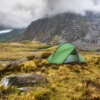 A lone green tent pitched on grassy, heather-studded moorland beside scattered boulders in a steep, rocky mountain valley partly shrouded in low clouds.