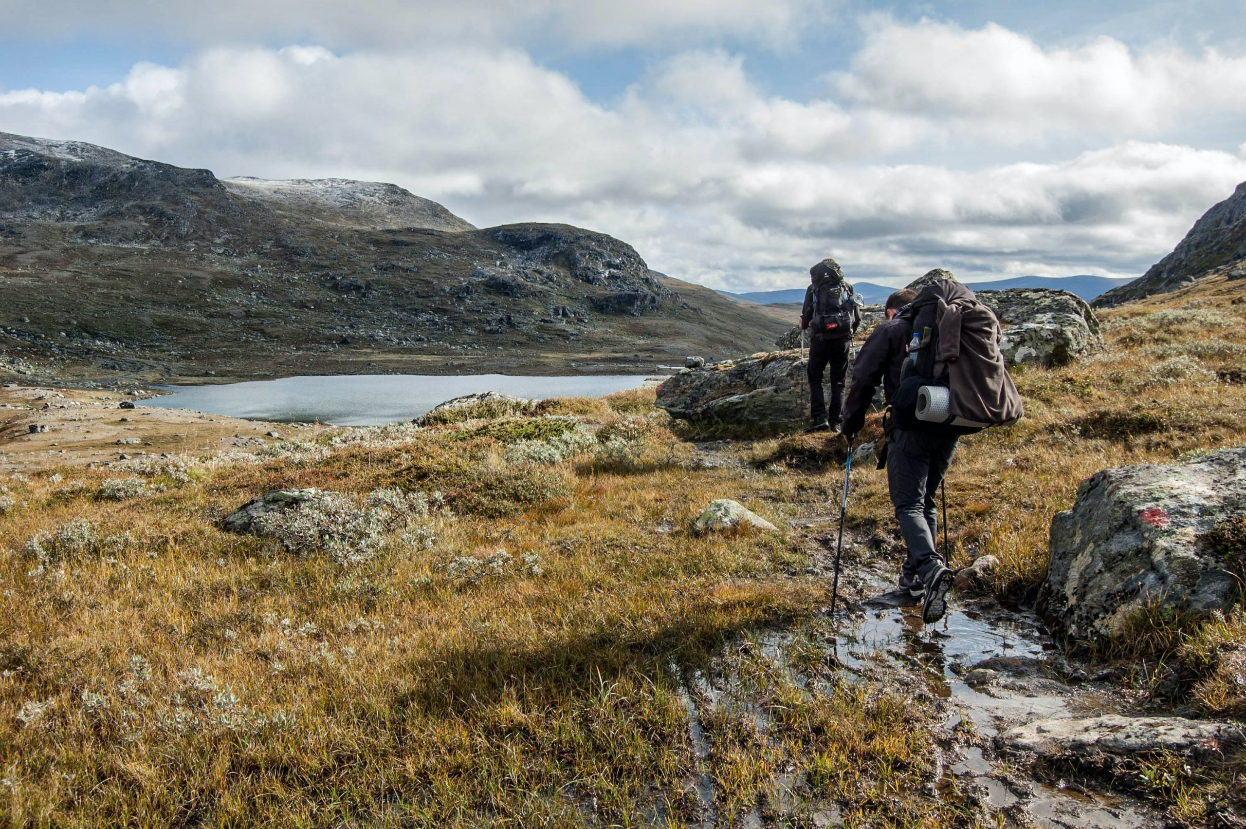 Two backpackers in dark outdoor gear using trekking poles walk a muddy trail past rocks and a small mountain tarn across a windswept grassy upland under a cloudy sky.