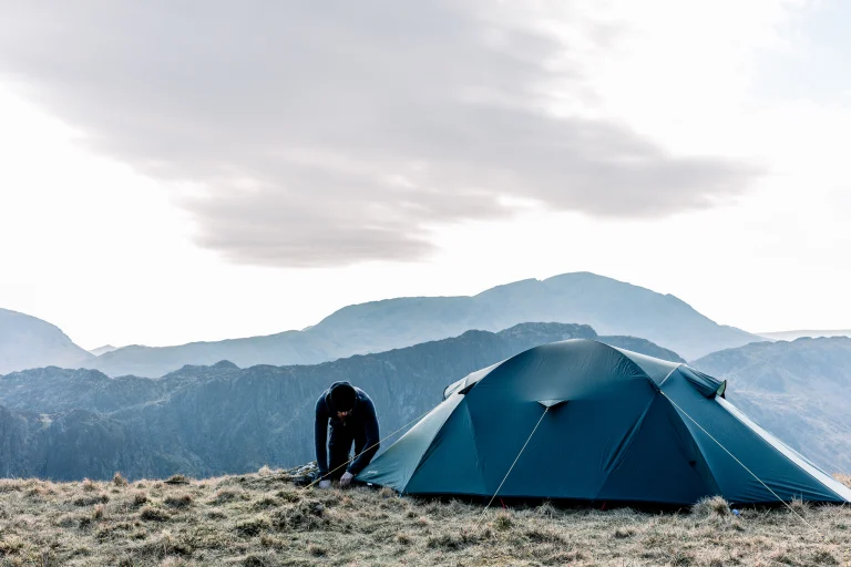 Person in dark clothing bending to secure a blue-green tent on a grassy mountain ridge with layered hills in the background under a pale, overcast sky