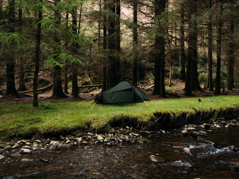 Green camping tent pitched on a grassy riverbank beside a rocky stream, set against a backdrop of tall, dense conifer trees.
