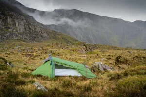 A small green tent with a partially open door and yellow and red guy lines pitched on grassy, rocky moorland beneath misty, steep mountain cliffs under an overcast sky.
