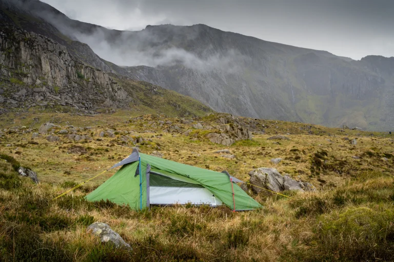 A small green tent with a partially open door and yellow and red guy lines pitched on grassy, rocky moorland beneath misty, steep mountain cliffs under an overcast sky.