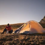 Camper in a red jacket sits on a grassy alpine plateau at sunset, unpacking gear from a foil container beside a pitched orange tent with rocky mountains in the background.