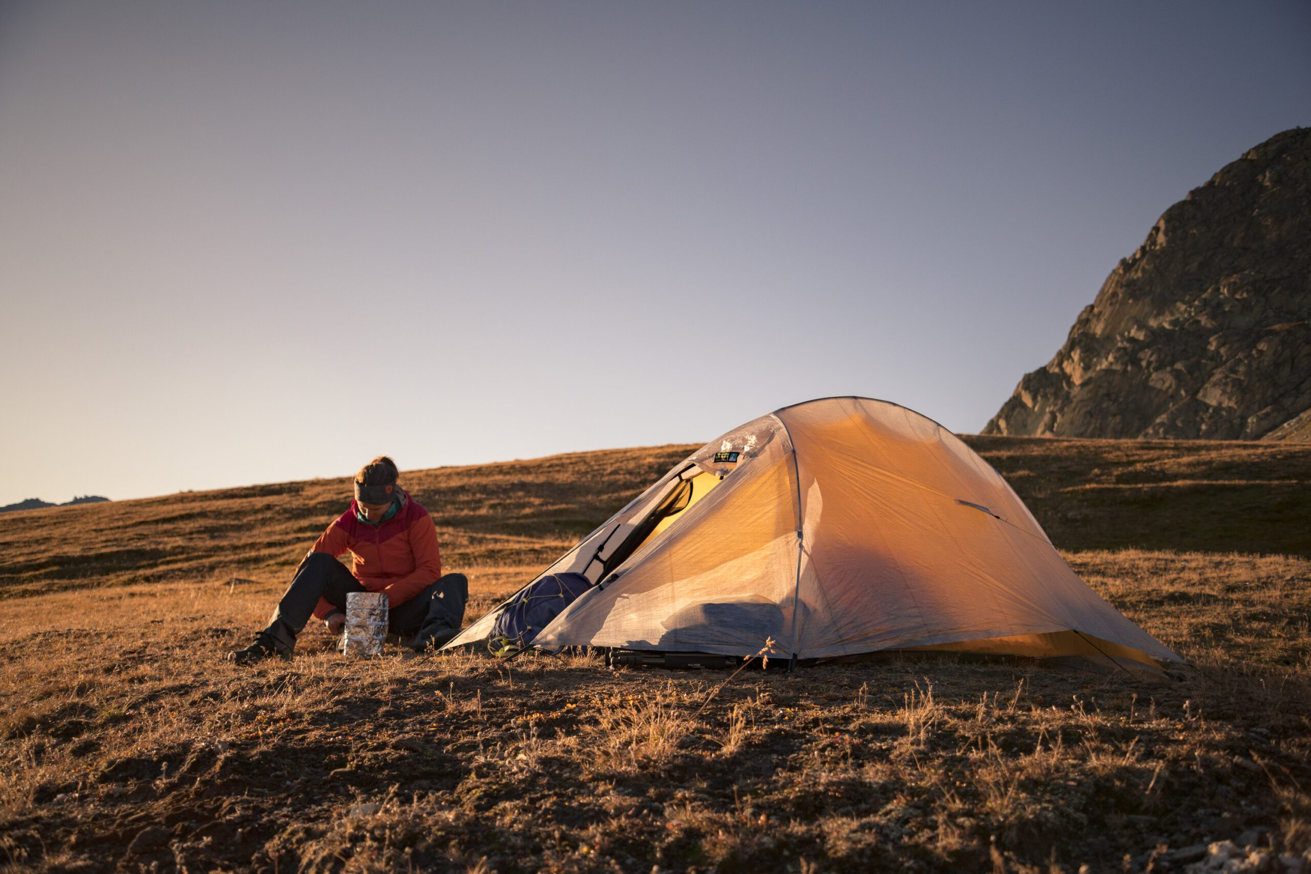 Camper in a red jacket sits on a grassy alpine plateau at sunset, unpacking gear from a foil container beside a pitched orange tent with rocky mountains in the background.