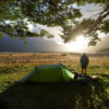 A green one‑person tent pitched under leafy tree branches on sunlit grass beside a wide river plain, a person standing near piled camping gear as the sun rises behind distant mountains.