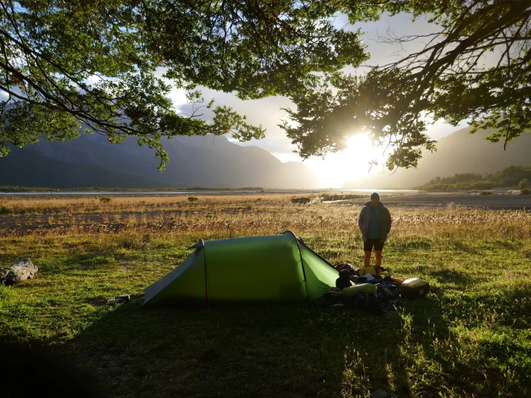 A green one‑person tent pitched under leafy tree branches on sunlit grass beside a wide river plain, a person standing near piled camping gear as the sun rises behind distant mountains.