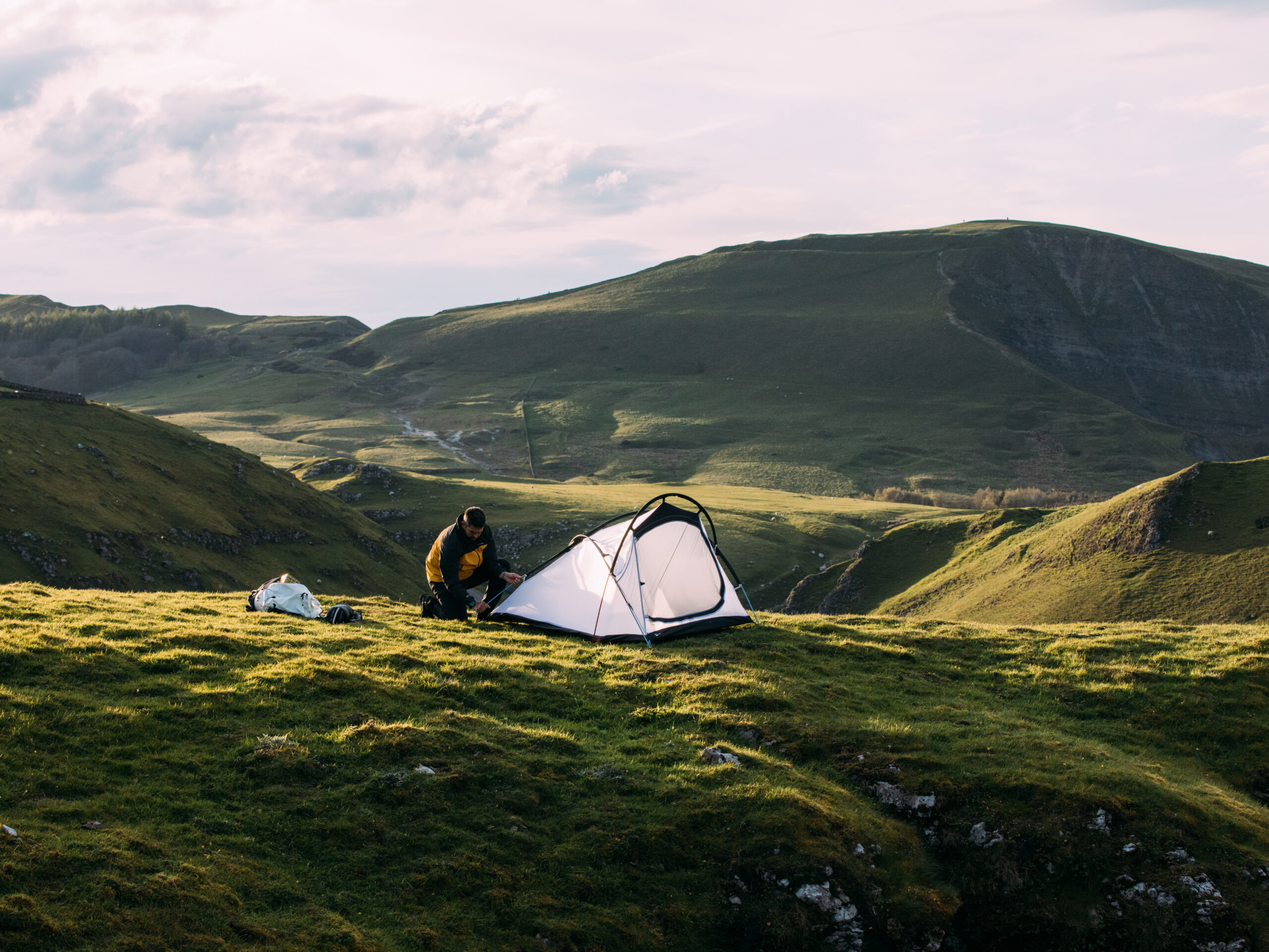 Person in a yellow-and-black jacket pitching a small white tent on a grassy ridge, with a backpack and gear nearby and rolling green hills under a pale sky in the background.