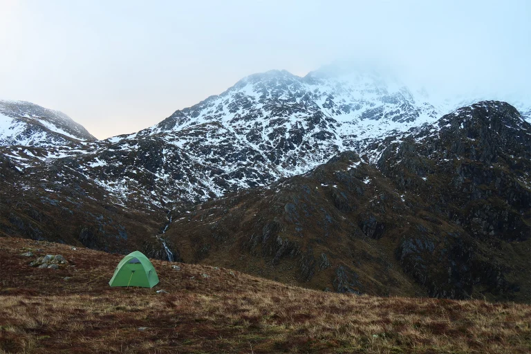 A solitary green tent on a brown grassy slope in the foreground with steep, snow-speckled rocky mountains and mist-shrouded peaks rising behind and a narrow stream running through the valley.