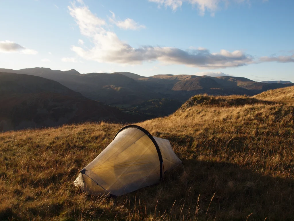 ls_tn_LASER-ULTRA-1-ANGLE-TARN-PIKES_2021 A small translucent backpacking tent pitched on a sunlit grassy hillside, overlooking rolling mountains under a blue sky with scattered clouds.