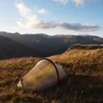 A small translucent backpacking tent pitched on a sunlit grassy hillside, overlooking rolling mountains under a blue sky with scattered clouds.