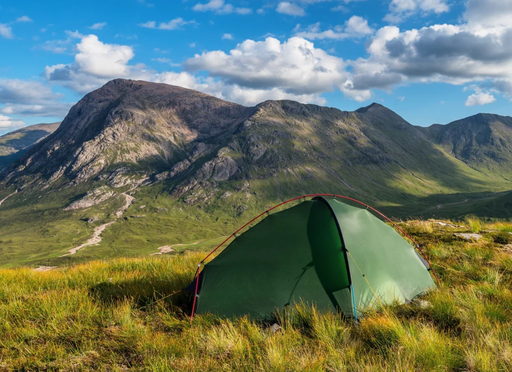 Green dome tent with red poles pitched on a grassy hillside in front of steep rocky mountains and a sunlit valley with a winding path under a blue sky dotted with white clouds