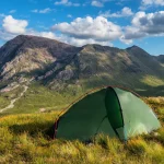 Green dome tent with red poles pitched on a grassy hillside in front of steep rocky mountains and a sunlit valley with a winding path under a blue sky dotted with white clouds