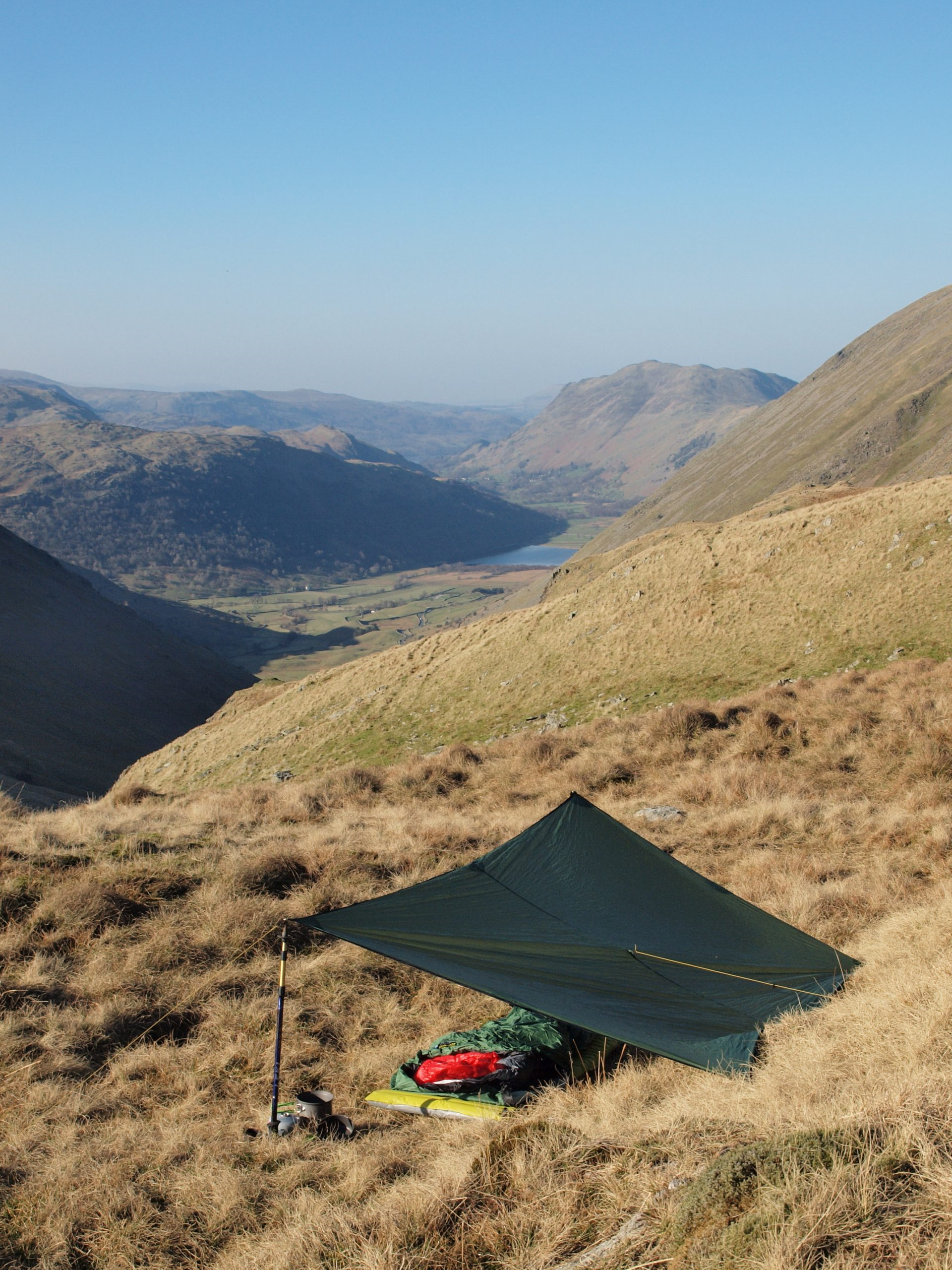 ls_tn_TARP-2_2021 Small green tarp shelter pitched on a windswept grassy hillside with a sleeping bag, mat and cooking gear by a trekking pole, overlooking a sunlit valley, distant lake and rugged hills under a clear blue sky.