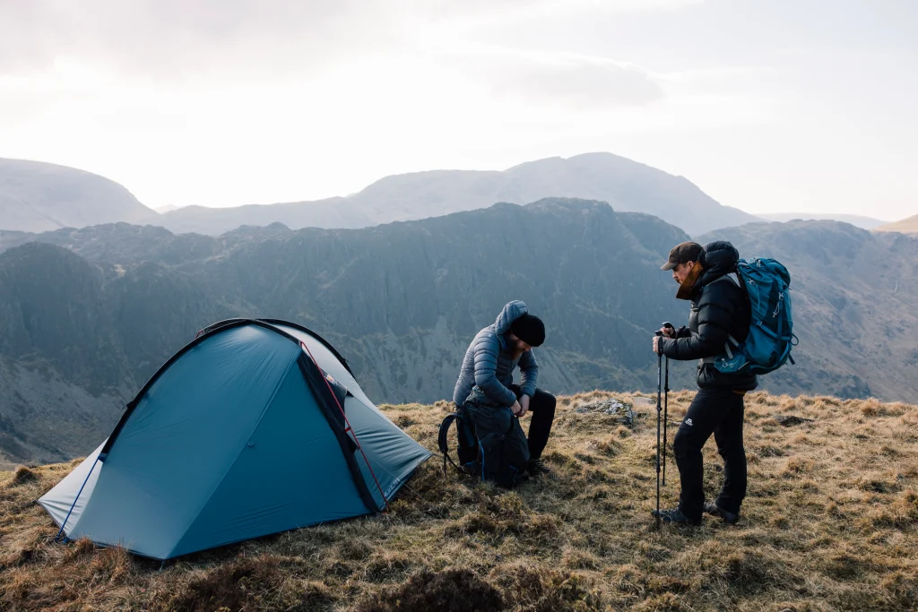 Two hikers on a grassy mountain ridge beside a small blue tent: one seated adjusting a rucksack, the other standing with trekking poles and a large blue backpack, with layered rocky peaks behind them.