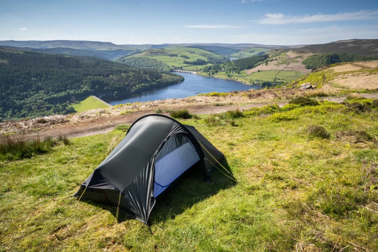 Small dark tunnel tent pitched on a grassy hillside overlooking a blue reservoir with a visible dam and patchwork green hills under a clear sky