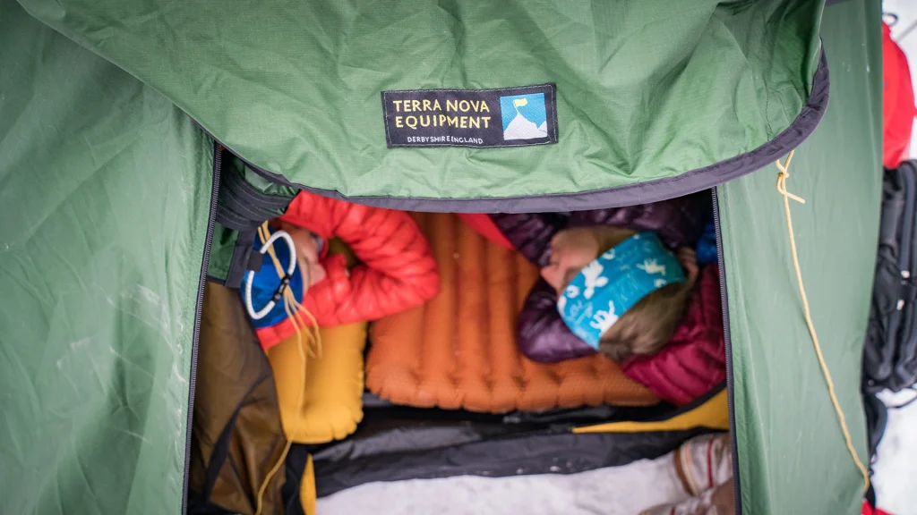 Top-down view into a green Terra Nova tent with its logo patch visible, showing two people in winter jackets lying on orange and yellow sleeping mats—one wearing a blue patterned headband—with snow visible outside.