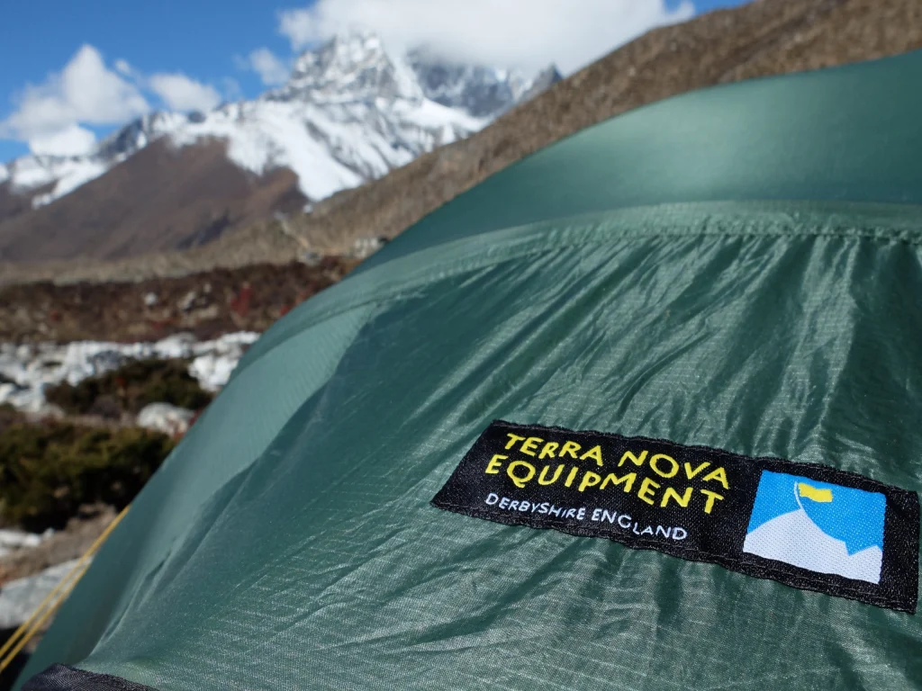 Green tent flysheet with a prominent 'Terra Nova Equipment, Derbyshire England' stitched label in the foreground and snow‑capped mountains and rocky alpine terrain blurred in the background.