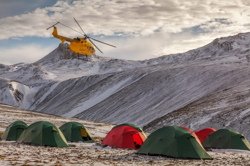 Yellow helicopter hovering above a cluster of green and red camping tents pitched on a snow-dusted alpine plain with steep, snow-covered mountain ridges under a cloudy sky.