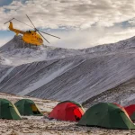 Yellow helicopter hovering above a cluster of green and red camping tents pitched on a snow-dusted alpine plain with steep, snow-covered mountain ridges under a cloudy sky.